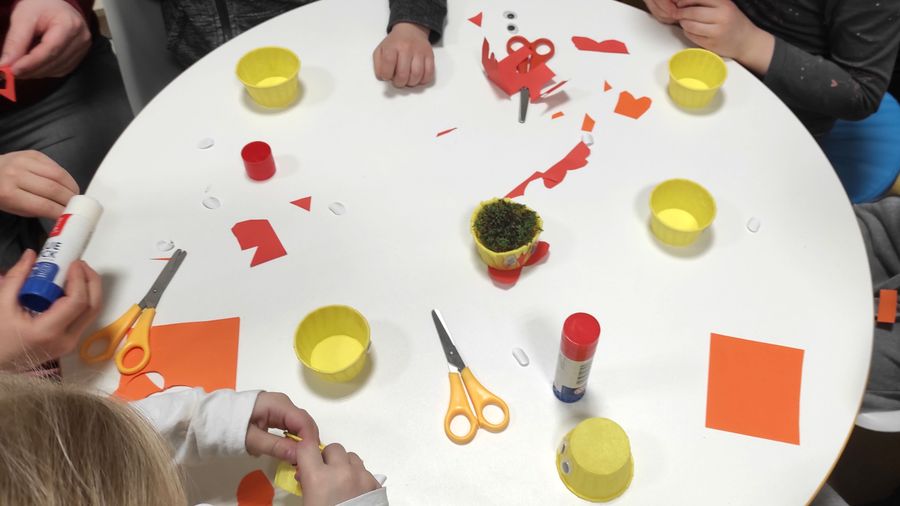 Dzieci rozbią pracę plastyczną. Zdjęcie przedstawia widok na stolik. Na stoliku leżą żółte pojemniki, papier kolorowy, nożyczki i klej, widoczne są dłonie dzieci.